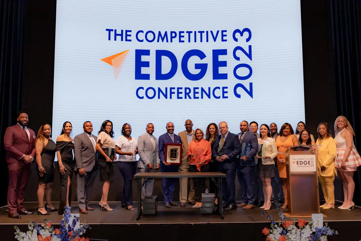 A group of people standing on stage in front of the competitive edge conference logo.